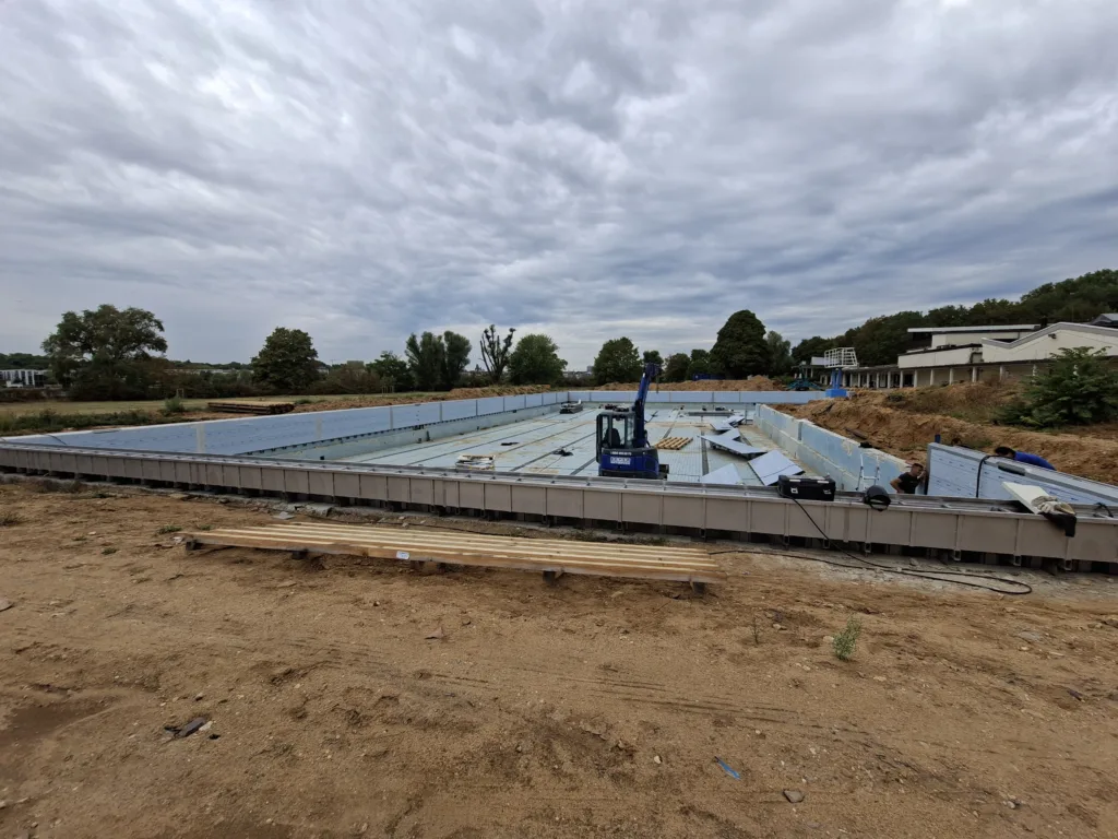 Baustelle eines großen Schwimmerbeckens mit hellblau ausgekleidetem Beckenboden und Beckenwänden. Im Becken stehen Baumaschinen und liegen Baumaterialien für die weitere Montage bereit.