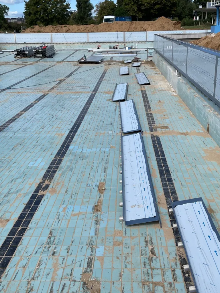 Blick in das Schwimmbecken während der Montage von Edelstahltafeln. Mehrere längliche Tafelelemente liegen auf dem gefliesten Beckenboden und werden von der Fußraste bis zum Boden eingebaut.