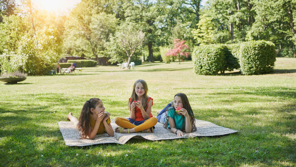 Drei Kinder essen Melone, sitzen auf deiner Decke im Gras